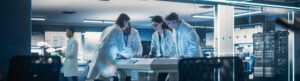 Four scientists in lab coats, likely scouted by top Life Sciences Recruiters, gather around a table in a modern laboratory. They engage in animated discussion, examining papers. The lab is brightly lit with advanced equipment and computer servers visible in the background.