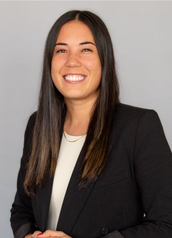 A person with long dark hair smiles, wearing a black blazer over a light-colored top, stands against a plain background.