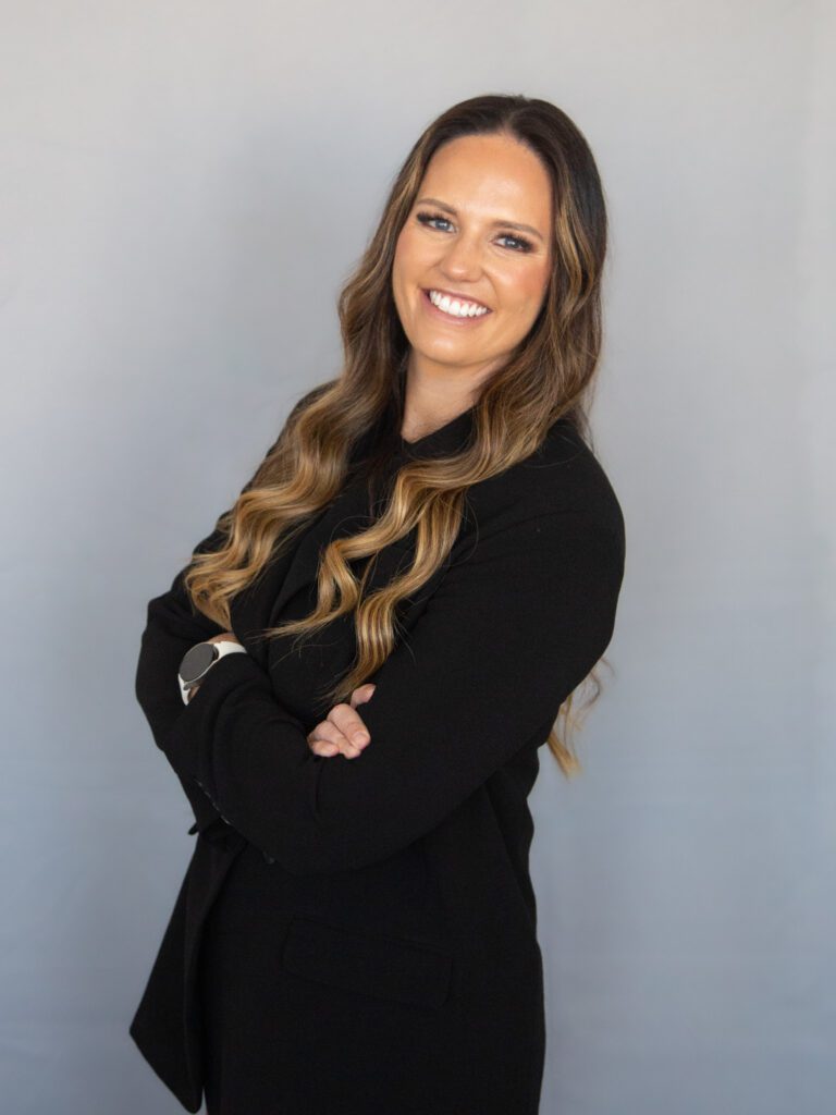 A woman with long wavy hair smiles, standing with her arms crossed. She is wearing a black blazer, a light watch, and is posed against a plain gray background.