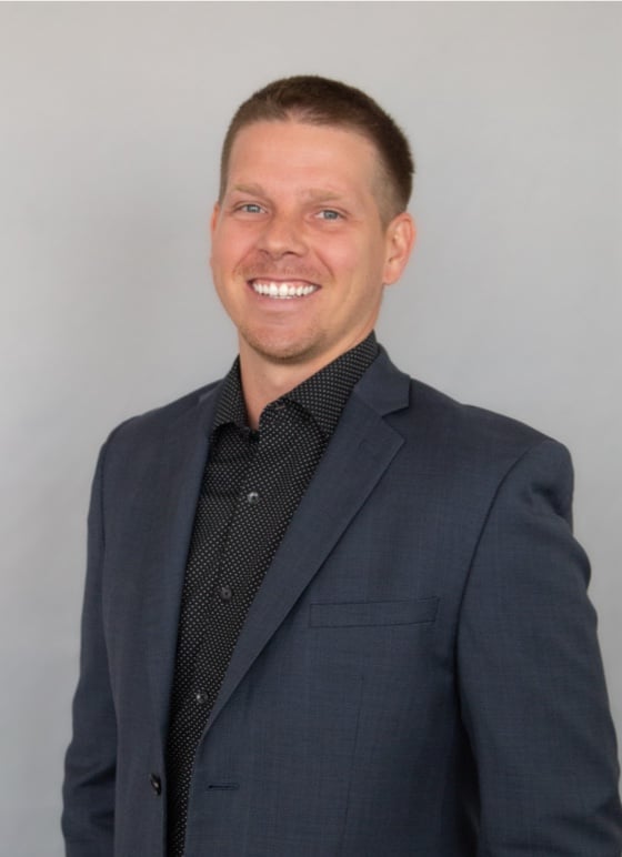 Smiling person in a dark suit and polka dot shirt, standing against a plain gray background.